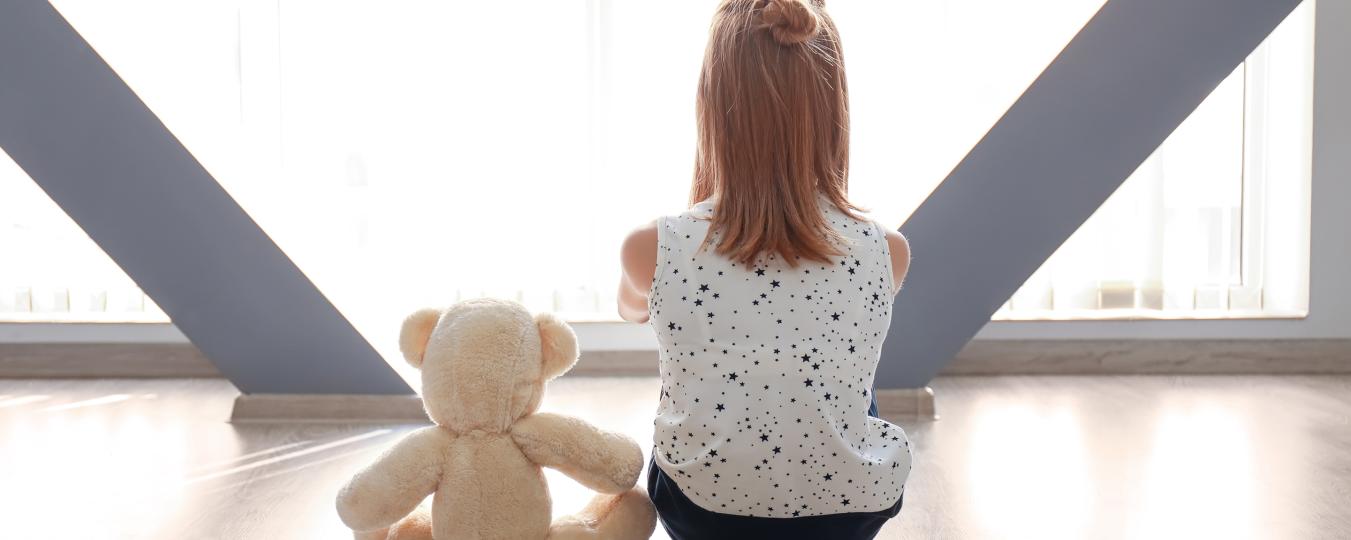 A child sits beside a teddy bear in front of a window