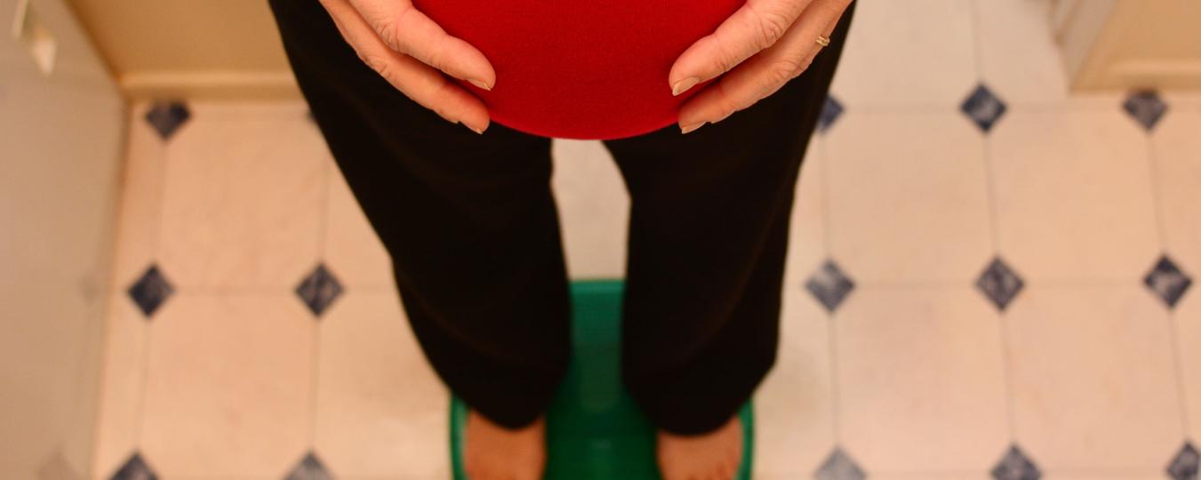 A pregnant woman cradles her bump while standing on a scale