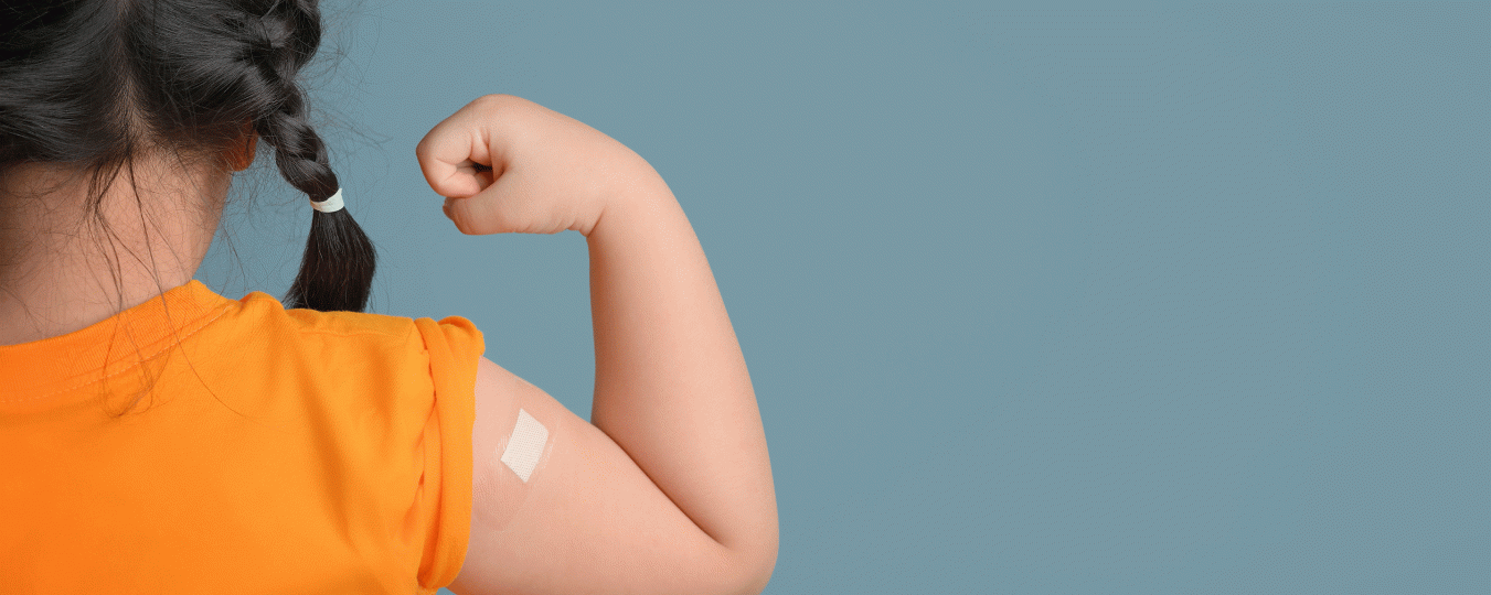 A child with french braids wearing an orange shirt holds up her arm to show off her vaccine bandaid.