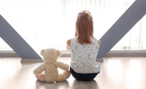 A child sits beside a teddy bear in front of a window