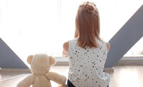 A young girl sits with a teddy bear in front of a window 