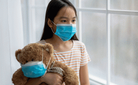 A child wearing a mask hugs a teddy bear, also wearing a mask