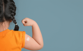 A child with french braids wearing an orange shirt holds up her arm to show off her vaccine bandaid.